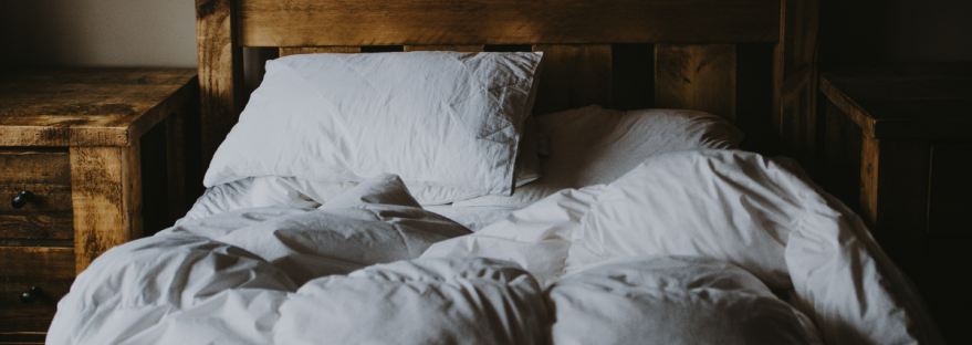 A bed with disheveled white sheets and a wooden, rustic nightstand
